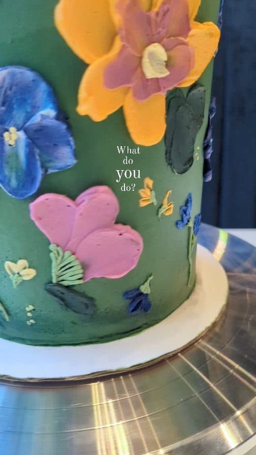 Still from a reel: a round cake painted with loose pink, blue, and green palette-knife flowers on white buttercream.
