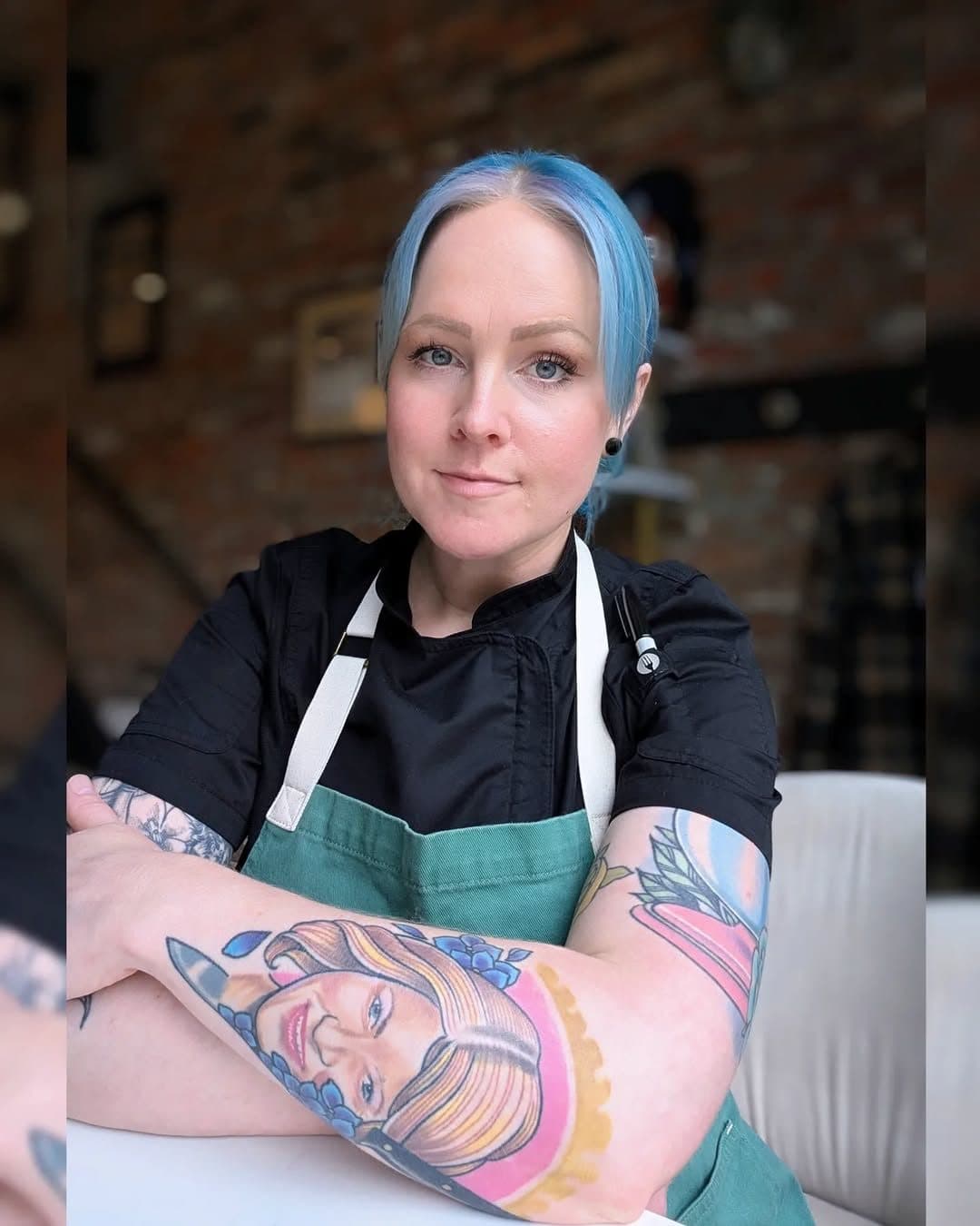 Rebecca Kelley in a commercial kitchen, blue hair and tattoos, arms crossed, wearing a black apron that reads Baking.