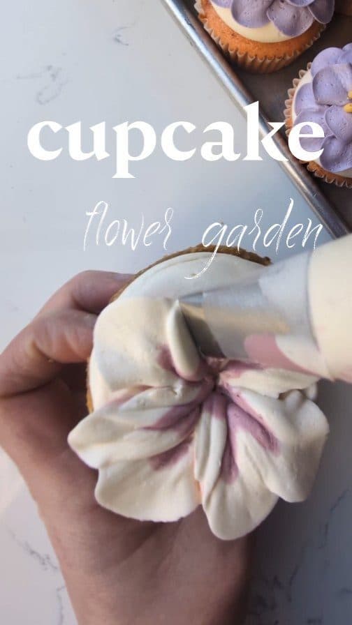 Still from a reel: a hand holding a piping bag, filling a bouquet of cupcakes with white, pink, and blue buttercream flowers.