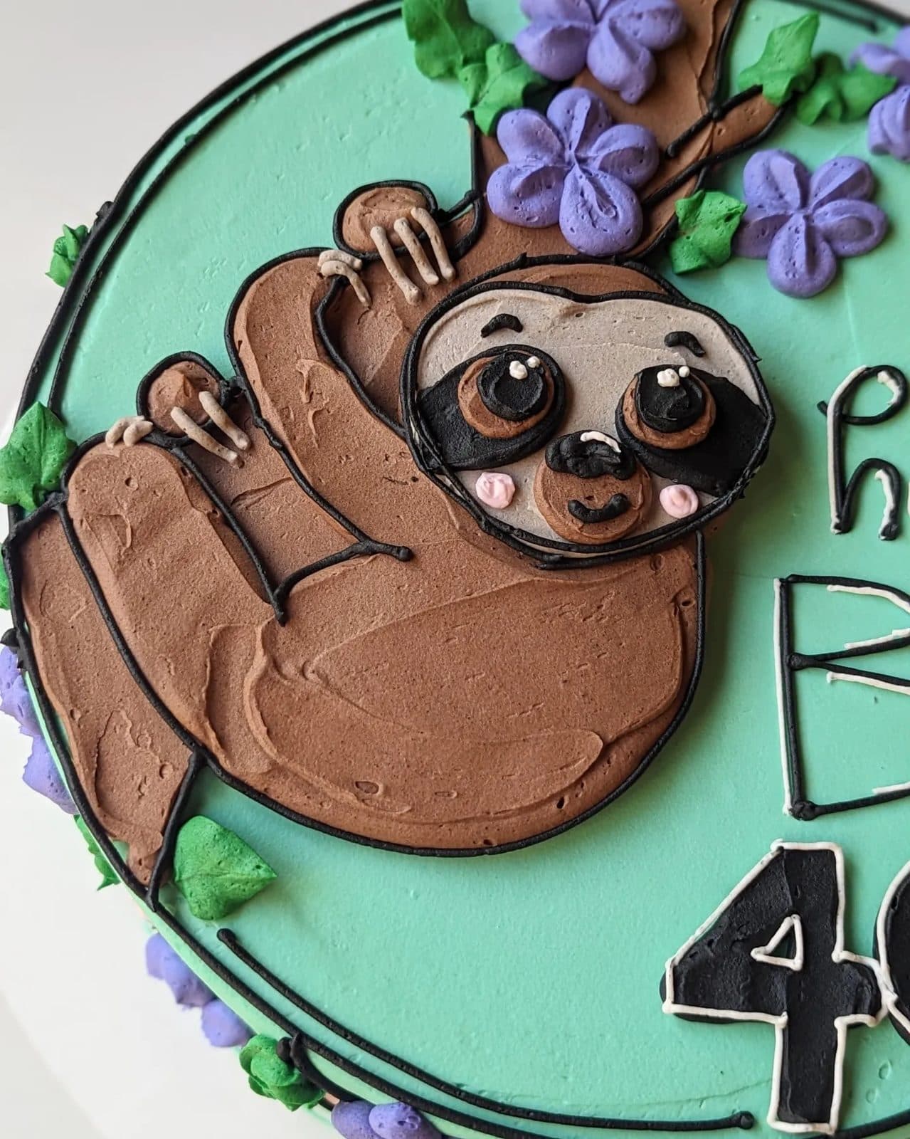 Closer view of the buttercream sloth's face, dark eye masks, and smile beside the green tree.